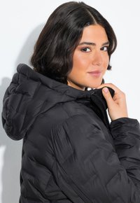 Černá péřová bunda s prošívaným designem, vybavená kapucí a stojáčkem. Materiál vypadá hladce a je zateplený.