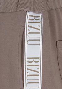 Brązowe dresy mają biały pasek po bokach z nadrukiem "BIZUU" w kolorze złotym. Tkanina jest miękka, z żebrowanym paskiem w talii i boczną kieszenią.