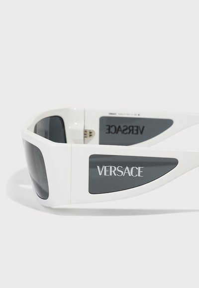 Hvite plastsolbriller med vinkelaktig design, gråtonede linser og "VERSACE"-logo på armen. Teksturert grå aksent på siden.