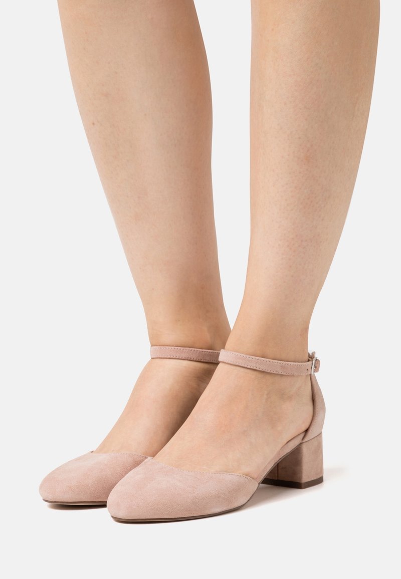 Pieds chaussés de chaussures à talons blocs en suède rose clair avec bride à la cheville sur fond blanc.
