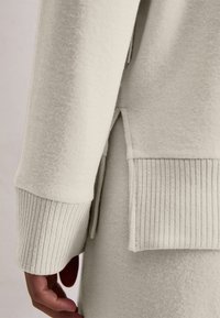 Gros plan sur une main à côté d'une manche et d'un ourlet de pull en tricot beige clair, avec des poignets côtelés et une petite fente sur le côté.