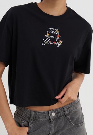 Person trägt ein schwarzes, bauchfreies T-Shirt mit weißer Schrift "Take care of Yourself" und kleiner bunter Blumenstickerei auf der Brust.