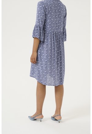 Femme portant une robe bleue fleurie longueur genou avec manches trois-quarts à volants et des chaussures slingback bleu clair à talons bas, debout de dos.