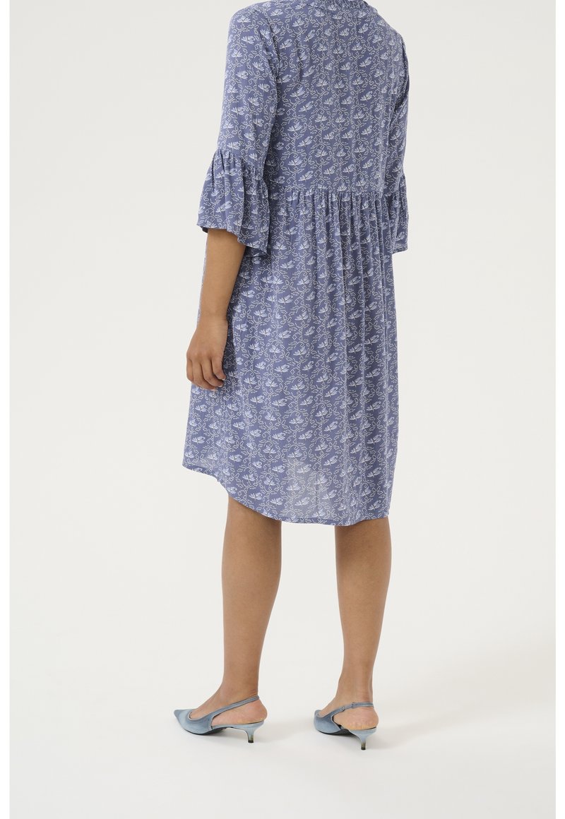 Femme portant une robe bleue fleurie longueur genou avec manches trois-quarts à volants et des chaussures slingback bleu clair à talons bas, debout de dos.