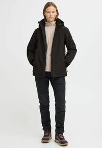 Svart vanntett jakke med hette, glidelås, sidelommer, brukt over en lys beige fleecegenser; kombinert med svarte bukser og tursko.
