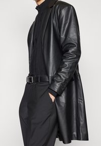 Czarny skórzany płaszcz o dopasowanym kroju, długiej długości i mankietach na guziki, noszony na czarnym dzianinowym topie i czarnych spodniach, z eleganckim paskiem.