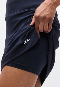 Μπλε σκούρα φούστα με εφαρμοστή γραμμή, ελαστικό ύφασμα και μικρή λευκή λεπτομέρεια με λογότυπο στο τελείωμα. Ο πολυεπίπεδος σχεδιασμός αποκαλύπτει ένα εσωτερικό σορτς.