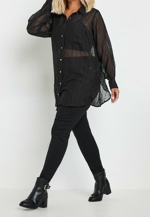Frau trägt eine transparente schwarze Bluse mit Knöpfen, schwarze Hose und schwarze Stiefeletten mit Schnalle, läuft vor einem schlichten weißen Hintergrund.