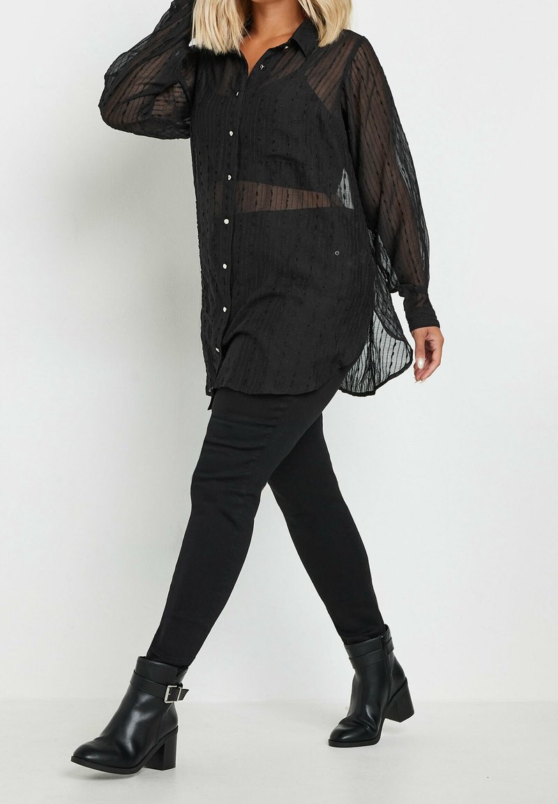 Femme portant un chemisier noir transparent à boutons, un pantalon noir et des bottines noires avec boucle, marchant devant un fond blanc uni.