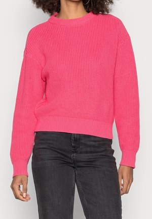 Osoba ubrana w jaskraworóżowy sweter z dzianiny z długimi rękawami i czarne jeansy, stojąca na tle jednolitego, jasnego tła.