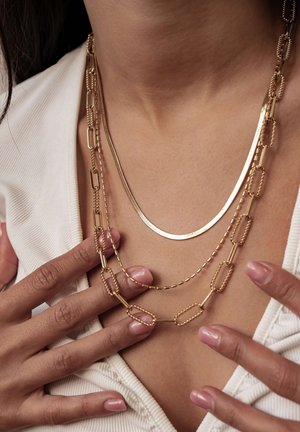 Brust einer Frau, geschmückt mit drei übereinander getragenen goldenen Halsketten, deren Finger mit glänzend nude-rosa lackierten Nägeln den Kragenbereich eines weißen Oberteils berühren.