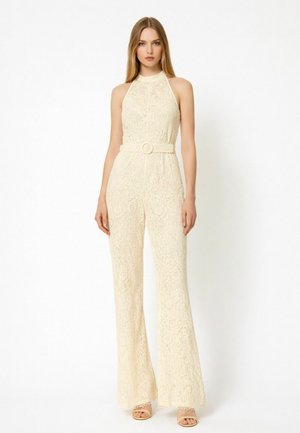 Donna bionda in piedi che indossa una tuta smanicata in pizzo panna con cintura di stoffa e gambe larghe, abbinata a tacchi aperti color nude.