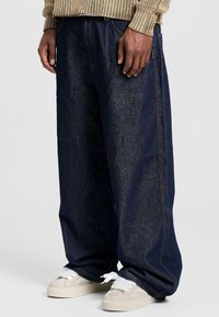 Pantaloni a gamba larga in denim blu scuro con cuciture a contrasto. Indossati con sneakers beige con dettagli bianchi e tessuto testurizzato.