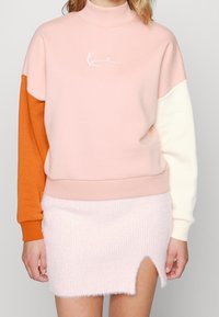 Sudadera a blocchi di colore in rosa chiaro, arancione e crema, con collo alto e logo ricamato; abbinata a una gonna morbida rosa con uno spacco laterale.