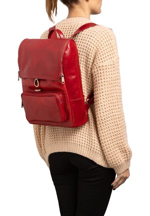 Femme portant un pull texturé beige et un pantalon noir avec un grand sac à dos en cuir rouge comportant plusieurs compartiments zippés.