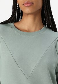 Sudadera de algodón verde con cuello redondo, costuras de hombro holgadas y un detalle de costura diagonal en el frontal. Textura suave.