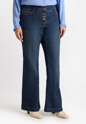 Jeans in denim blu scuro con gamba larga, chiusura frontale a cinque bottoni e dettagli di cucitura sottili. Abbinati a stivali alla caviglia beige.