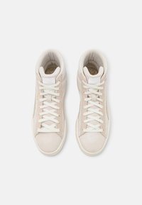 Paire de baskets montantes en daim beige clair avec des lacets blancs et des semelles en caoutchouc, vues de dessus sur fond blanc.
