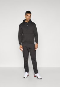 Un sweat à capuche gris foncé avec une poche avant et un cordon, associé à un jogging assorti avec des coutures courbées. Des chaussures de sport blanches avec des accents rouges.