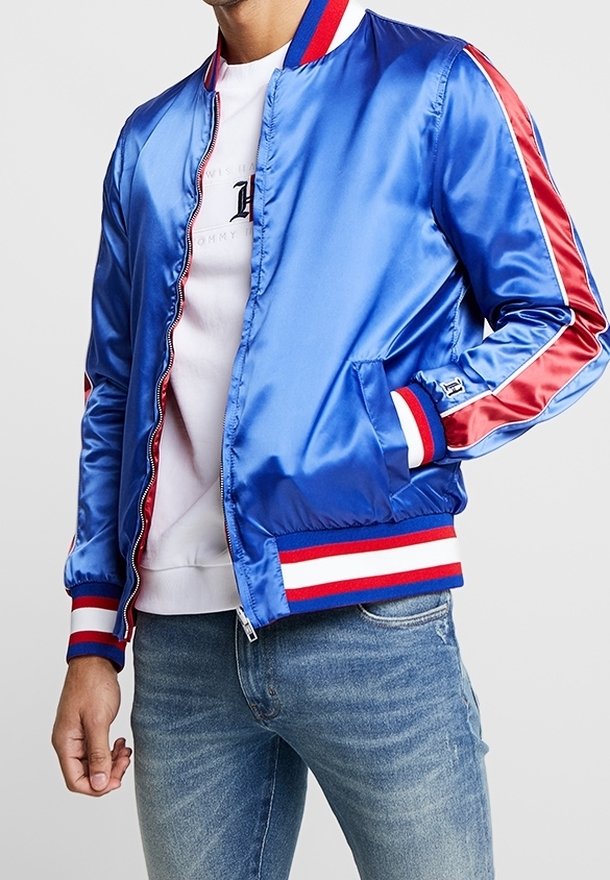Homme portant une veste bomber bleue brillante avec des poignets et des finitions rayés rouges et blancs, une chemise blanche et un jean bleu.