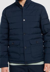 Marinblå puffjacka med ståkrage, knappstängning, sidofickor och horisontellt quiltad struktur för isolering.