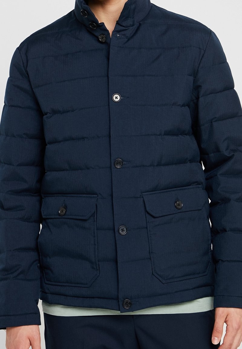 Marinblå puffjacka med ståkrage, knappstängning, sidofickor och horisontellt quiltad struktur för isolering.