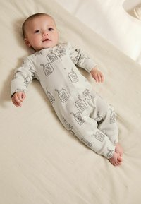 Krämfärgad babydräkt med lång ärm, prydd med ett svart juiceförpackningsmönster och knäppning, utlagd på en strukturerad ljus tygyta.