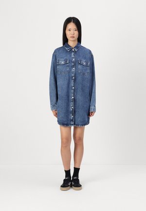 Denim shirt dress, light blue, button-up with two chest pockets, long sleeves, and a straight cut, featuring a hem with slight fraying details.