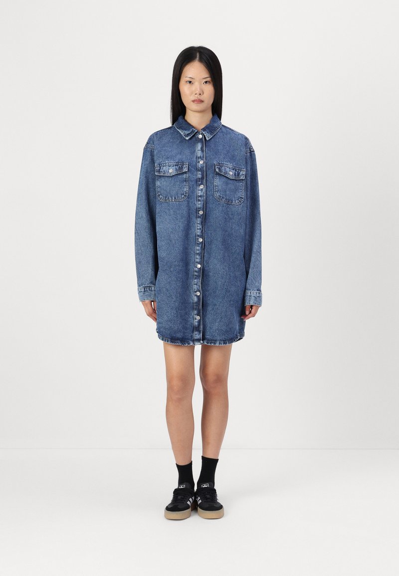 Vestito camicia di denim, azzurro chiaro, con bottoni e due tasche sul petto, maniche lunghe e taglio dritto, con un orlo con lievi dettagli sfilacciati.