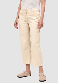 Pantalones de pierna amplia en color crema, fabricados en algodón. Presentan dos bolsillos frontales, cierre de botón y un ajuste relajado. Combinados con zuecos beige.