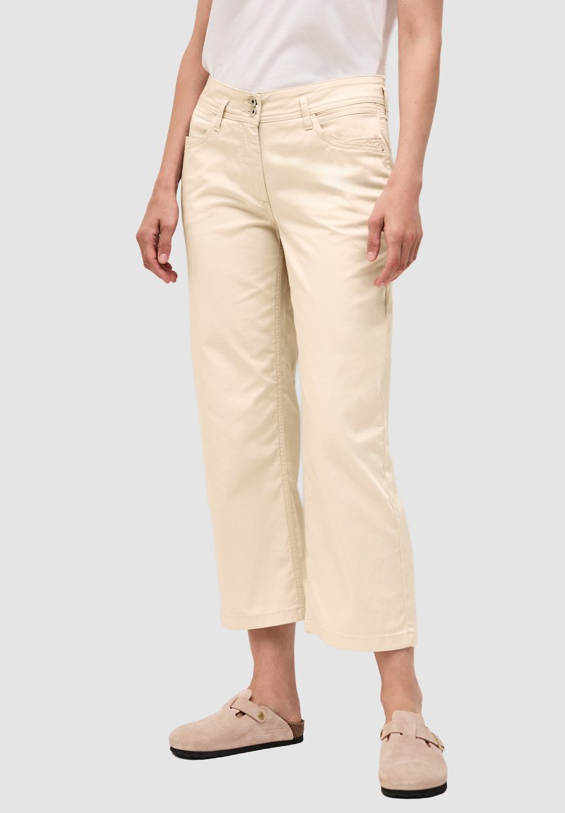 Pantalones de pierna amplia en color crema, fabricados en algodón. Presentan dos bolsillos frontales, cierre de botón y un ajuste relajado. Combinados con zuecos beige.