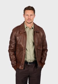 Hnědá kožená bunda se zipem vpředu, dvěma předními kapsami a žebrovanými manžetami, nošená přes světle zelenou košili na knoflíky a tmavé kalhoty.