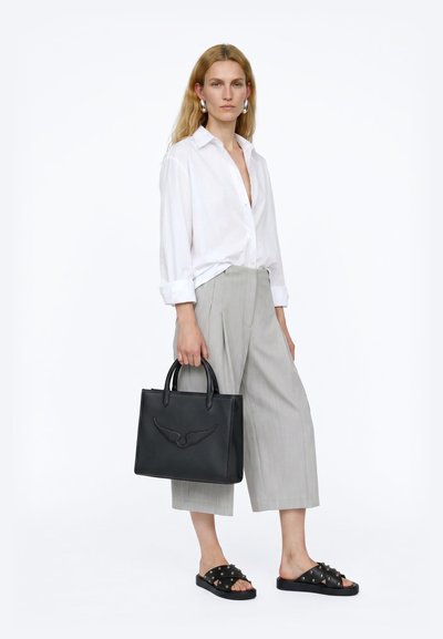 Femme portant une chemise blanche et un pantalon gris à jambes larges, portant un sac à main noir et des sandales à clous noires.