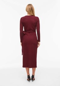 Robe en maille bordeaux à manches longues, corsage ajusté, avec une ceinture à nouer à la taille, s'arrêtant à mi-mollet. Chaussures à talons hauts noires.