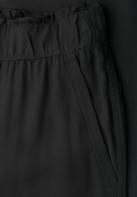 Close-up van zwarte broek met een elastische tailleband en een schuine zijzak op een donkere achtergrond.