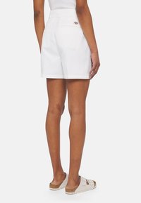 Shorts bianchi in cotone con una vestibilità dritta, tasche posteriori e un piccolo logo vicino all'elastico in vita; indossati con sandali di colore chiaro.