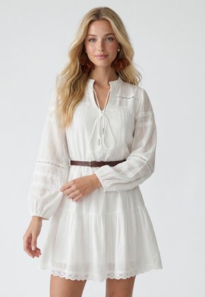 Donna con lunghi capelli biondi che indossa un vestito bianco con bordo in pizzo, cintura marrone e grandi orecchini marroni a fiori, in piedi davanti a uno sfondo semplice.