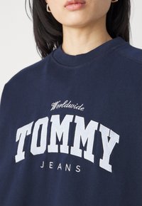 Felpa blu navy con scollatura a girocollo a costine; "TOMMY" in lettere bianche, "Worldwide" sopra e "JEANS" sotto in carattere più piccolo. Tessuto morbido.