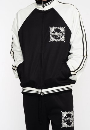 Veste de survêtement zippée noire et blanche et pantalon avec logos floraux blancs et texte sur la poitrine et la cuisse, modèle les mains dans les poches.