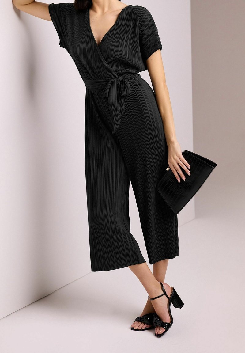 Femme portant une combinaison noire plissée avec une taille nouée, des sandales noires à talons et tenant une pochette noire.