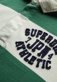 Groen en wit gestreepte poloshirt met marineblauwe geborduurde tekst "SUPER JPN ATHLETIC" op de borst. Katoenen materiaal, met gestructureerd detail.