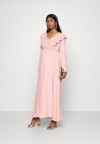 Longo vestido rosa feito de tecido suave. Apresenta detalhes de babados nas mangas e uma cintura atada. Combinado com acessórios dourados e sandálias de salto.