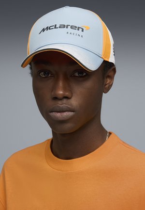 Ung person iført en lyseblå og orange McLaren Racing-kasket og orange crewneck-trøje, der vender fremad mod en grå baggrund.