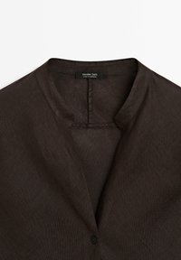 Chemise en lin brun foncé avec col mandarin, un seul bouton noir et des détails de coutures près de l'encolure.