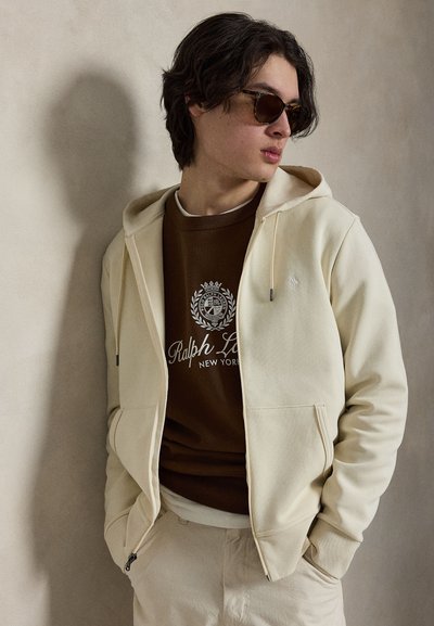 Νέος άνδρας που φοράει καφέ φούτερ Ralph Lauren, μπεζ φούτερ με φερμουάρ, παντελόνια στο ίδιο χρώμα και γυαλιά ηλίου, στέκεται δίπλα σε έναν απλό τοίχο.