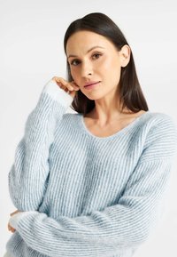Jasnoniebieski sweter w paski, wykonany z miękkiego, puszystego materiału, z dekoltem w serek i prążkowaną teksturą. Posiada długie rękawy z kontrastowymi mankietami.