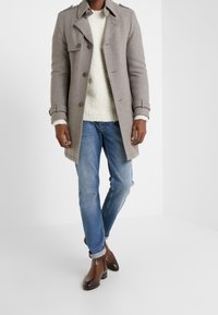 Grå uld trenchcoat med dobbeltbrystet design, knapdetaljer, en tekstureret cremefarvet sweater, blå jeans og brune ankelstøvler.