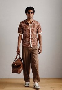 Brown patterned short-sleeve shirt with white accents, paired with beige trousers and white sneakers. A brown leather bag is held in one hand.