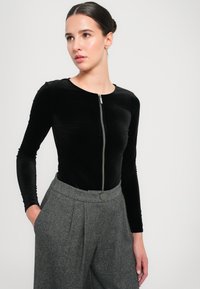 Čierny sametový body s dlhými rukávmi a okrúhlym výstrihom, so zipsom vpredu, v kombinácii s širokými nohavicami v šedom károvanom vzore, ktoré majú detail gombíka.