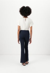 Haut blanc à manches courtes avec un détail en trou de serrure au dos, associé à un pantalon évasé bleu marine, porté avec des tongs noires. Design simple et ajusté.